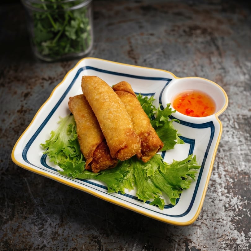 Three golden-brown spring rolls on a bed of lettuce, with a side of sweet chili sauce.