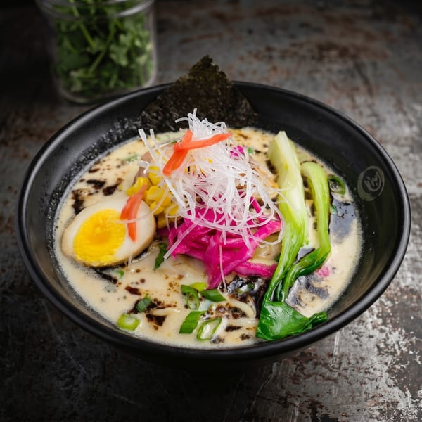 Close-up of a ramen bowl with creamy broth, egg, corn, bok choy, and pickled vegetables.
